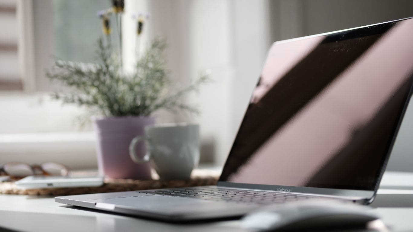 Imagem de um notebook desligado, ao fundo um vaso com flores e uma caneca de porcelana